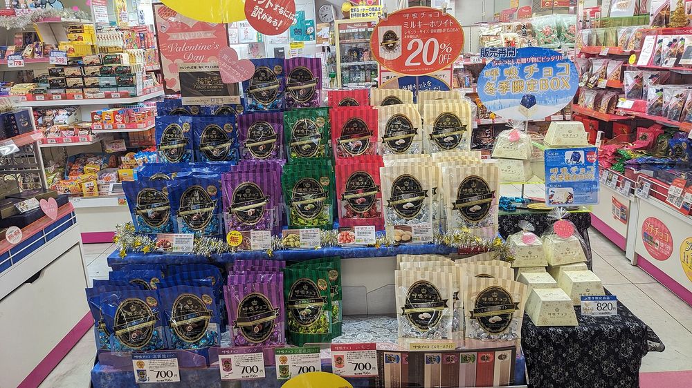 日本地下街糖果店的情人節專區:藍、紫、綠、紅、米色的巧克力包裝袋堆成階梯狀,每一款前面插著 700 円的手寫價卡,上方掛著粉紅色 HAPPY Valentine's Day 看板與冬季限定 BOX 的圓形吊牌