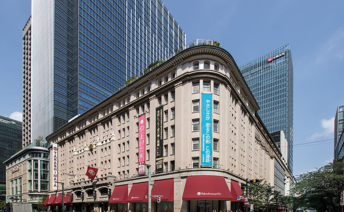 日本橋高島屋本館外觀:米色石造建築的轉角立面,一樓整排紅色遮陽棚印著 Takashimaya S.C.,左側垂著紅底白字 TAKASHIMAYA 直幅,上方外牆掛著粉紅與黑色的活動直幅