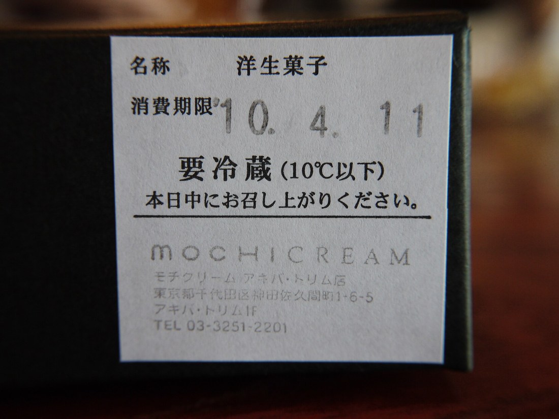 深色紙盒側面貼著一張白色標籤,由上而下印著「名称 洋生菓子」「消費期限 10. 4. 11」「要冷蔵(10℃以下)」「本日中にお召し上がりください。」,下方是店名與東京千代田區的地址電話