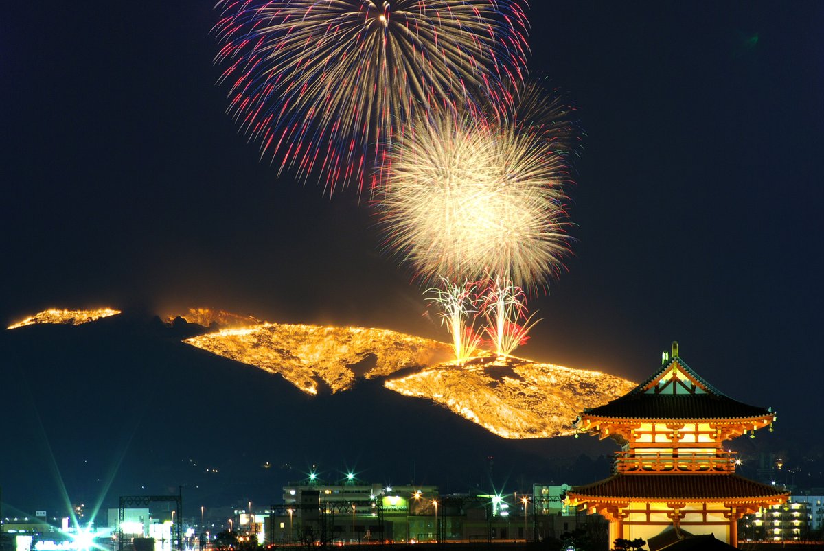 長時間曝光拍下的若草山燒山夜景：山坡上的火線連成一整片金黃色火面，上方同時炸開兩顆大花火，右下角是亮燈的平城宮跡朱雀門