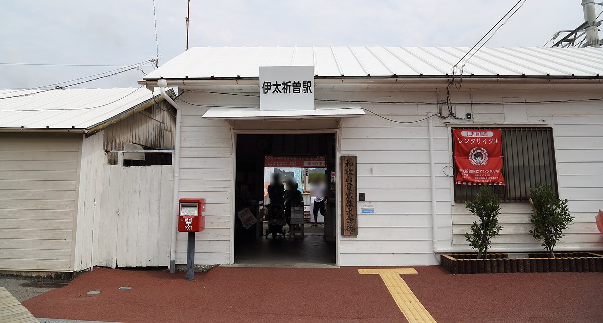 伊太祈曽站的白色木造站房:屋簷下白底黑字站名牌「伊太祈曽駅」,入口旁直掛木牌「和歌山電鐵株式会社」和紅色郵筒,右邊窗上掛著紅布條「たま自転車 レンタサイクル 伊太祈曽駅にてレンタル中」,地面是紅色鋪面和黃色導盲磚