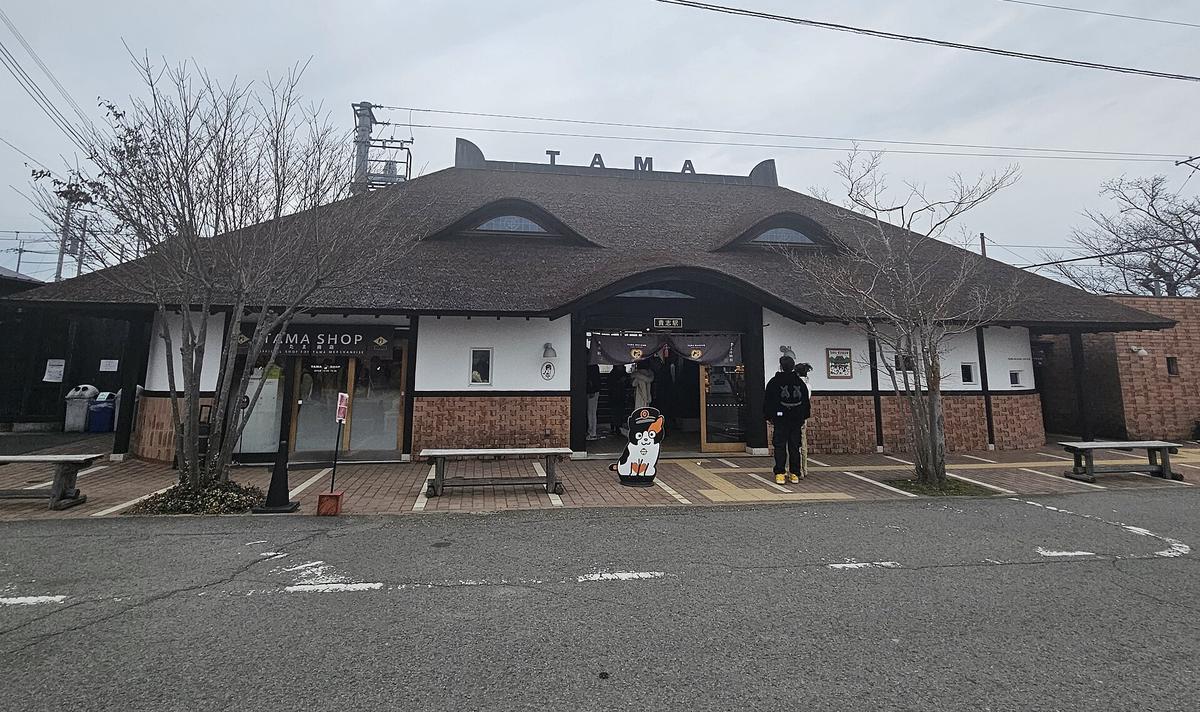 陰天冬季的貴志站站房正面:貓耳形屋脊和「TAMA」字樣、兩扇貓眼窗的大屋頂,中央入口上方是「貴志駅」站名牌與深色暖簾;左側一間店面招牌寫 TAMA SHOP,入口前有戴站長帽的三毛貓立牌、木頭長椅,兩位旅客背對鏡頭站在右側窗前