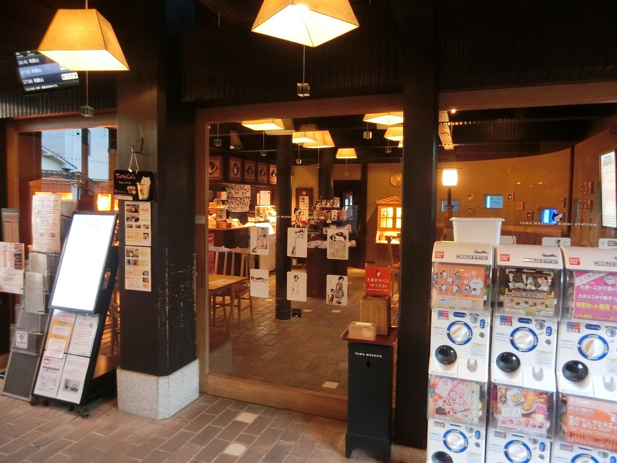 貴志站站房內たまカフェ的入口:深色木柱上掛著「TAMA CAFE」黑色小招牌,玻璃門裡是木桌椅、吊燈和貼著貓插畫的展示架;左邊立著菜單看板,右邊一排 BANDAI 扭蛋機,左上角牆上是發車時刻的電子看板