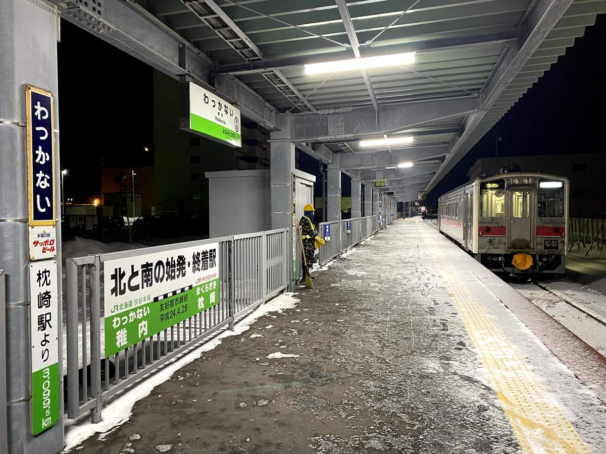 清晨天還沒亮的稚內站月台,屋簷下的地面覆著薄雪,一名戴黃色安全帽、整張臉包住的作業員拿著掃把,右邊停著一節紅白色的普通列車,左邊柱子上是「わっかない」站名牌與「北と南の始発・終着駅」看板