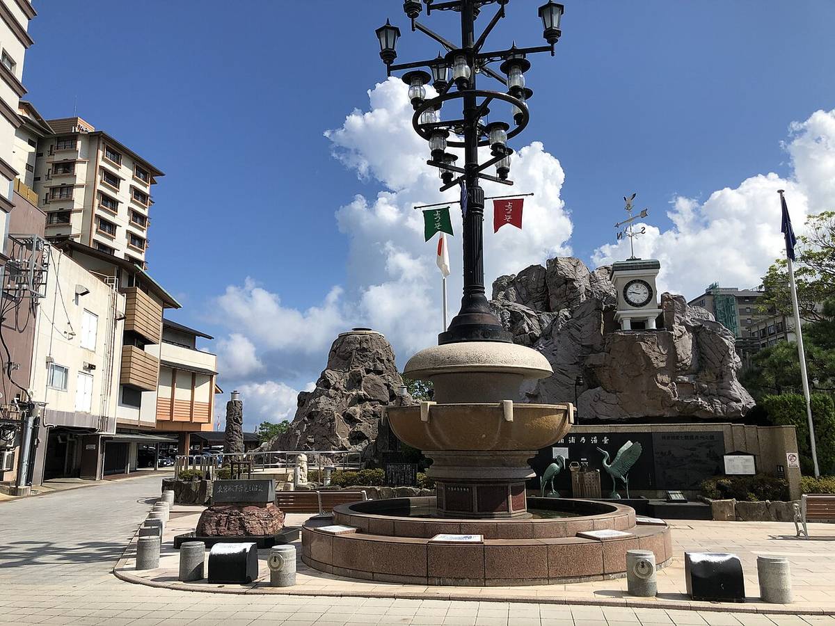 A public square in the Wakura Onsen town centre: a circular stone basin with a tall cast-iron lamp standard carrying several lanterns and two small welcome banners, artificial rockwork and a small white clock tower behind it, ordinary town buildings on either side, brick paving and nobody about