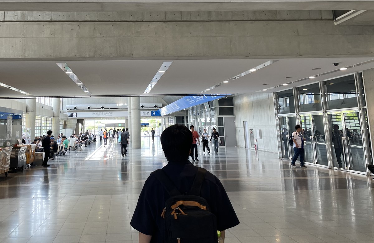 幕張展覽館館外的室內長廊:天花板下一條藍色長條印著館號 8,遠處白色指示牌寫著「東エントランス(ホテル方面)」,右側一排玻璃門,左側有人坐著休息,前景一人背對鏡頭