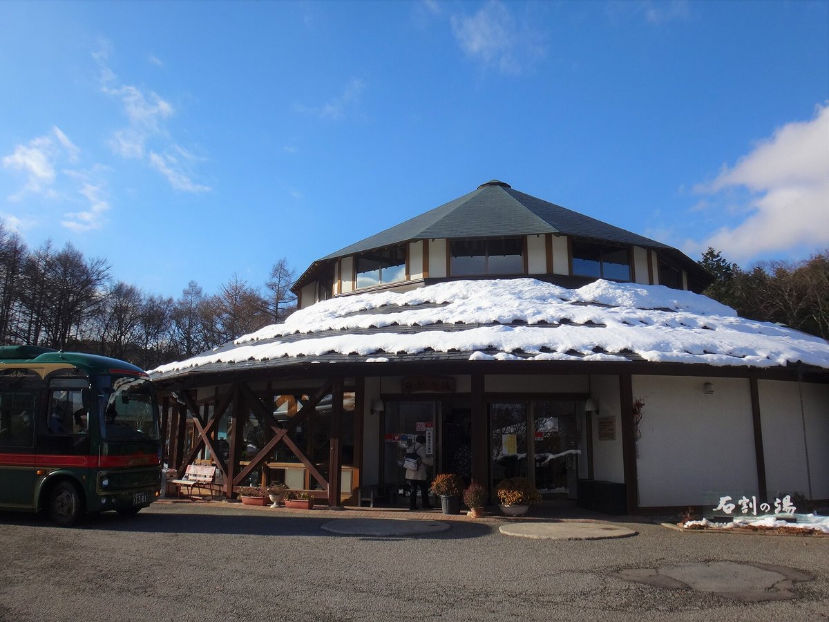 石割の湯的正面：八角形屋頂的溫泉建築，下層屋簷蓋著一圈積雪，右下角有「石割の湯」字樣，一位客人背對鏡頭站在入口，左邊停著一台墨綠色巴士