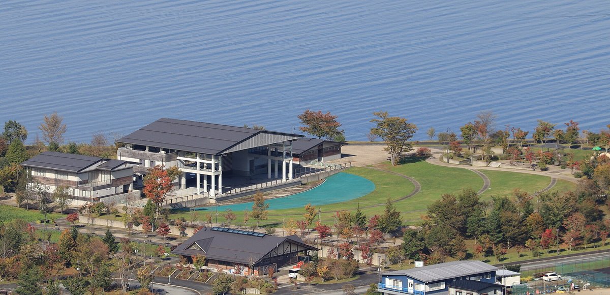 從山上俯瞰山中湖交流プラザきらら：湖邊幾棟深灰屋頂的建築、一片藍綠色的淺水池和弧形草坪步道，草坪外緣是淺色沙灘，上半部整片是湖面