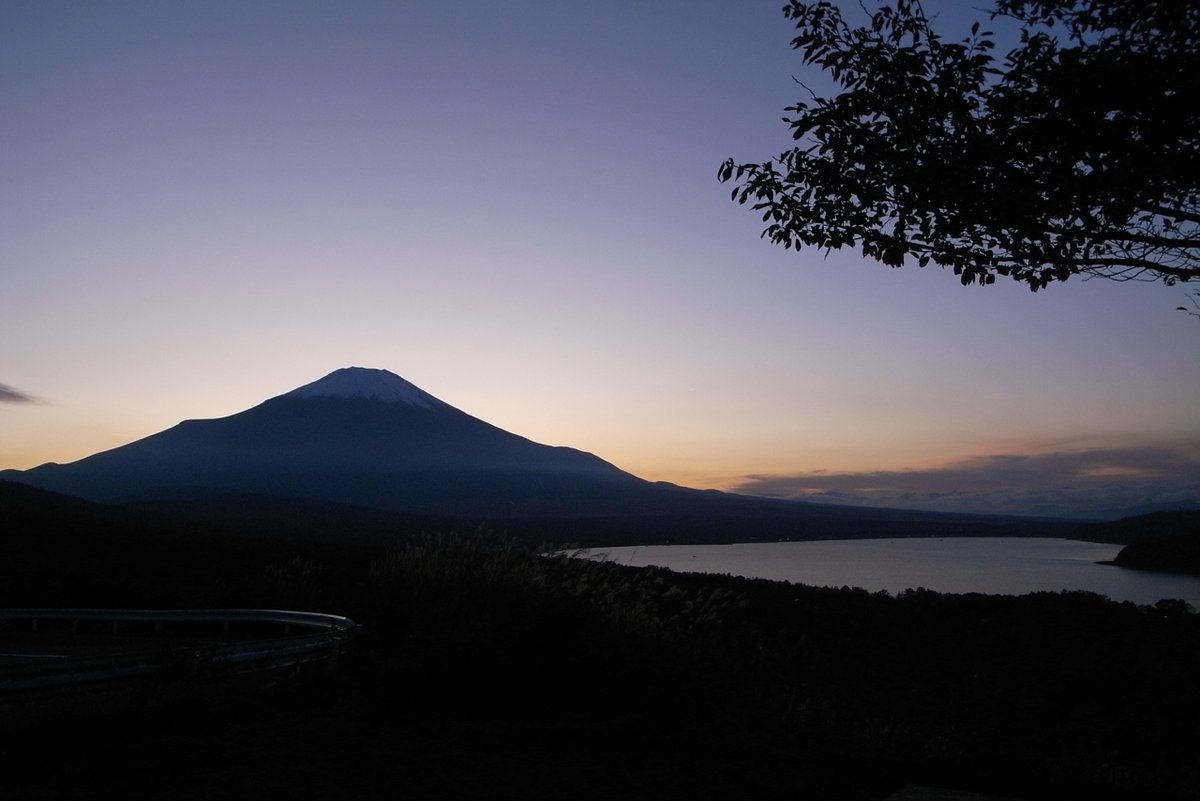 日落後從パノラマ台看山中湖：富士山成了深色剪影，地平線上一道橘色晚霞往上漸層成淡紫色天空，右下是暗下來的湖面，右上角有樹枝剪影