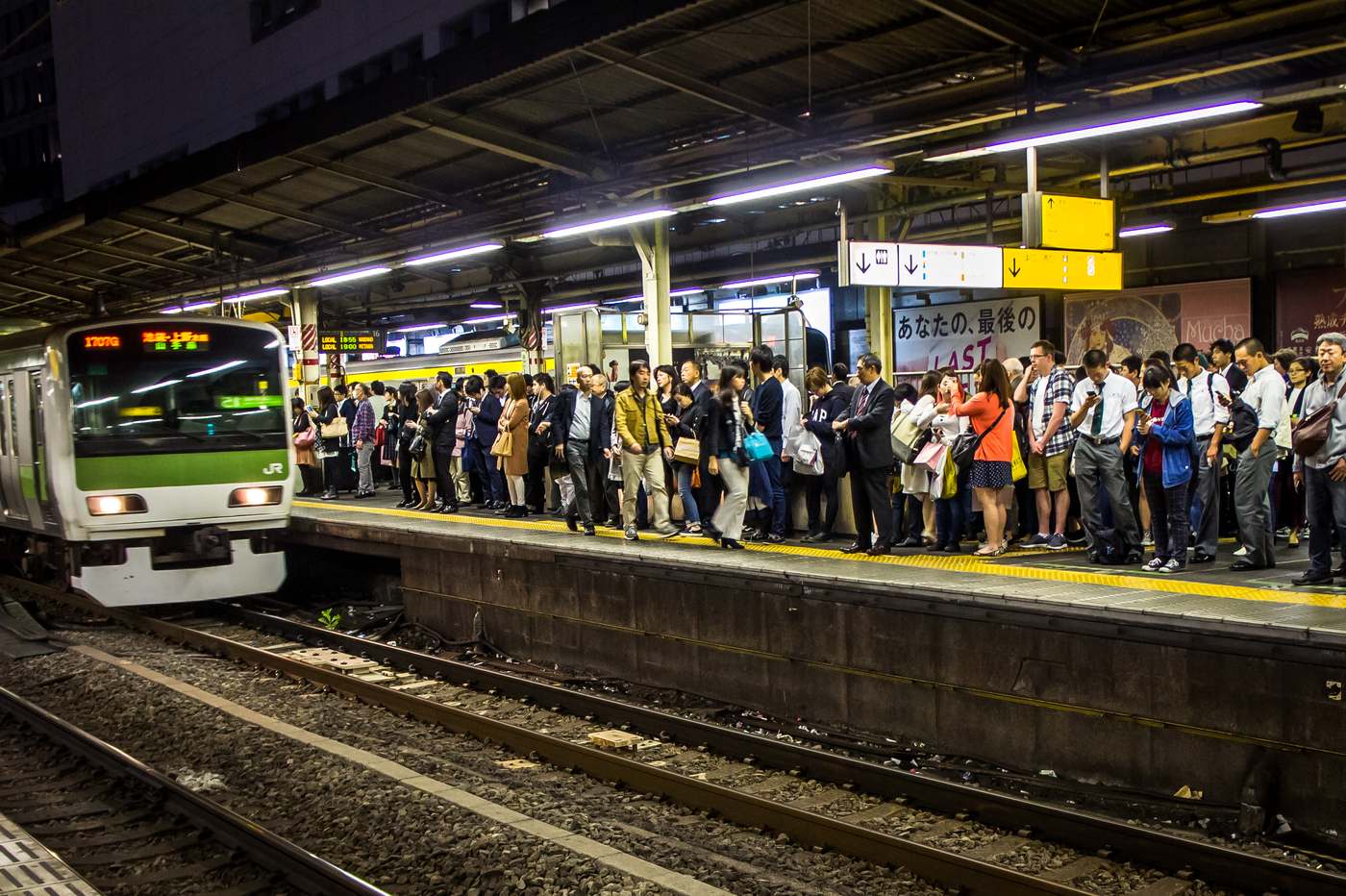 A Yamanote Line platform at night, packed with waiting passengers standing several deep along its whole length, while a green-banded train pulls in from the left with its headlights on; yellow tactile paving runs along the platform edge and yellow direction signs hang overhead.