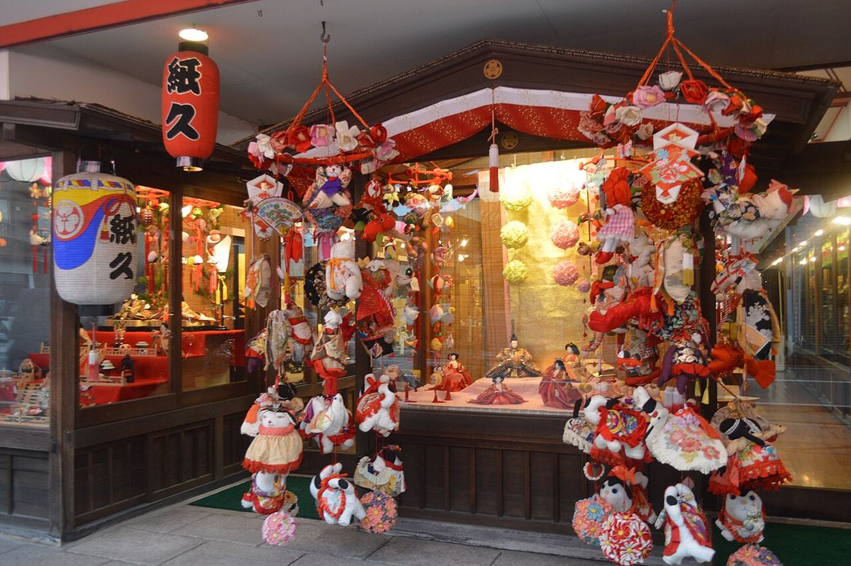 人形店「柳川紙久本店」的店門口:屋簷下掛著兩大串さげもん,吊的是穿紅色衣飾的布偶動物與大花鞠,後面櫥窗裡擺著雛人形,左邊掛著寫「紙久」的紅燈籠與白燈籠