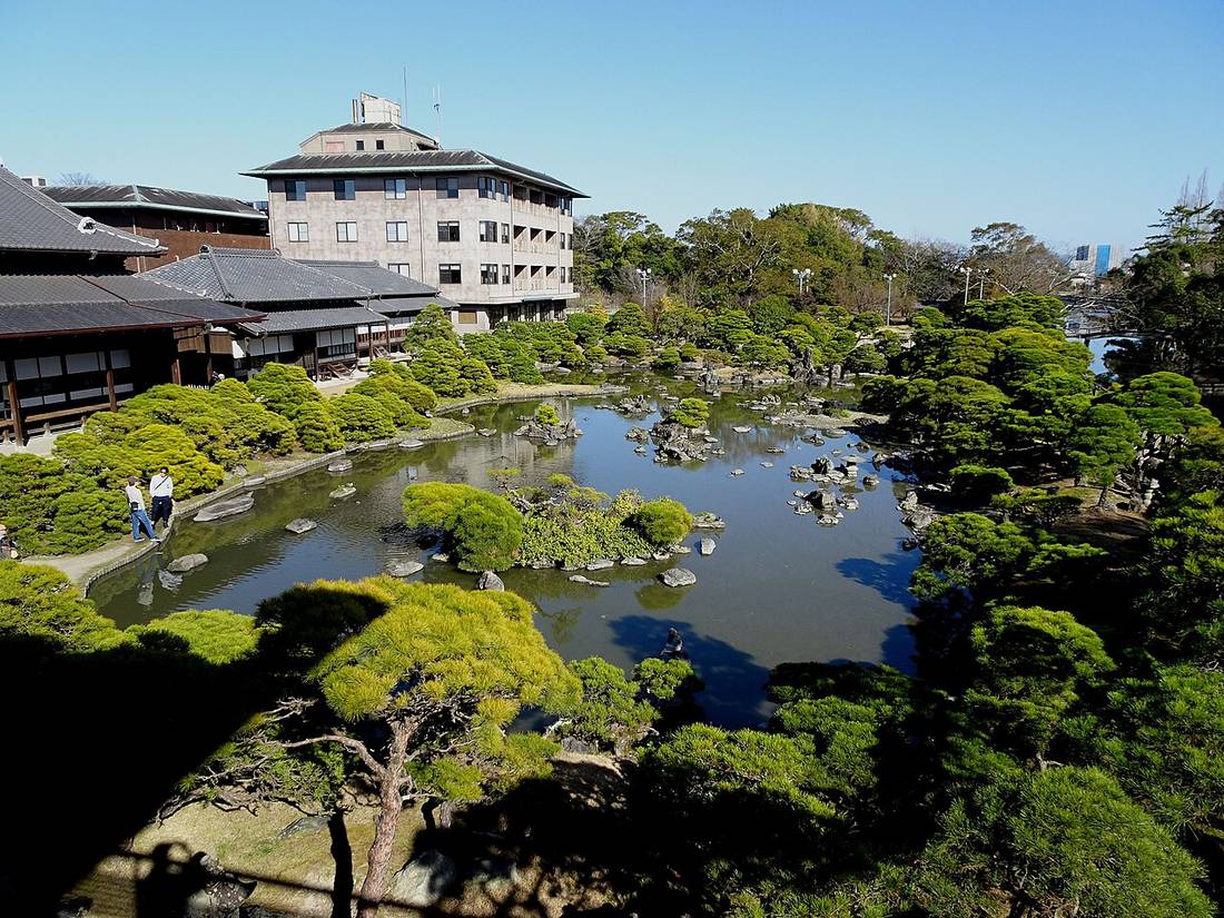 從高處看御花的庭園松濤園:池塘裡散布著岩石與小島,四周是修剪過的松樹,左邊是日式建築的瓦屋頂,後方一棟多層樓的建築,晴朗的藍天