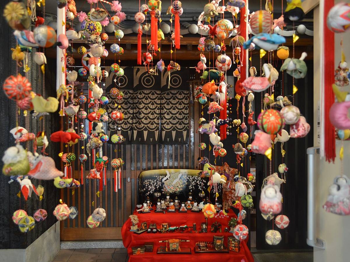 店內的雛祭陳設:下方鋪紅布的台子上擺著雛人形與小道具,後面立著畫有鶴的黑色屏風,上方與左右垂滿一串串さげもん,有刺繡的鞠、布製的兔子、鳥、金魚與紅白流蘇