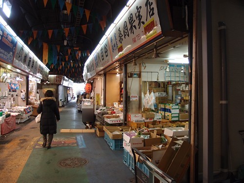 柳橋連合市場有頂棚的窄走道:左邊一位穿黑色長大衣的行人往裡走,右邊果實店門口堆滿紙箱與藍色塑膠箱,頭頂掛著一串串三角旗