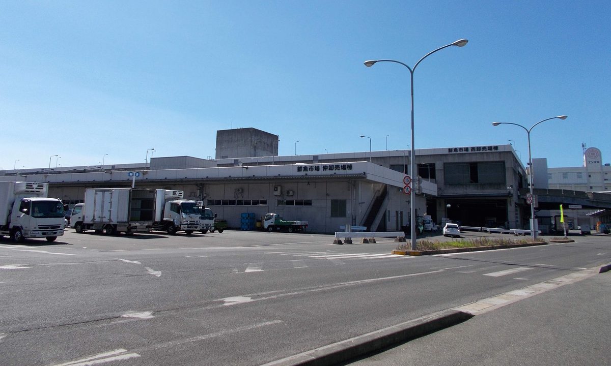 長濱的福岡市鮮魚市場:大片停車場後方是灰白色低層建築,外牆寫著「鮮魚市場 仲卸売場棟」與「鮮魚市場 西卸売場棟」,前面停著幾台冷藏貨車
