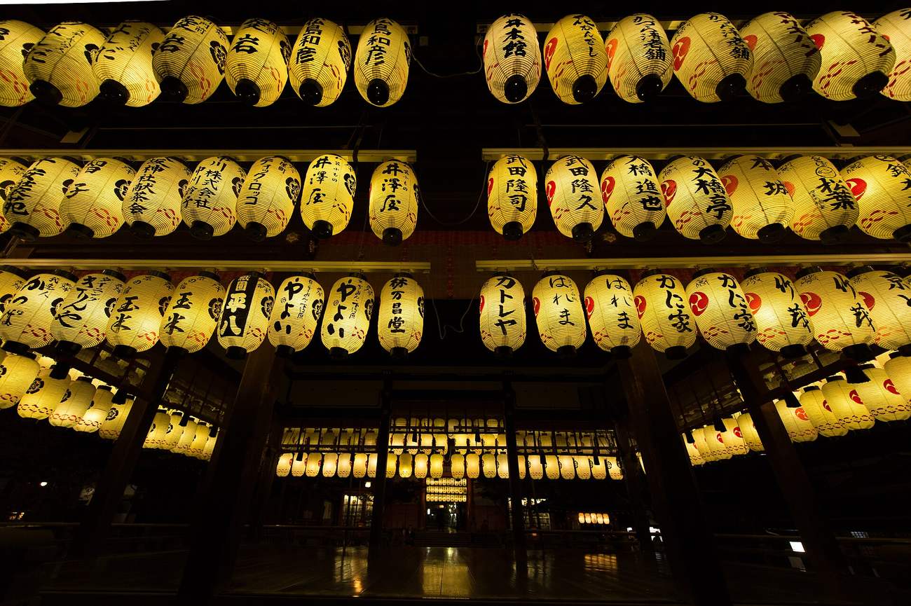Rows of lit white paper lanterns hanging from the Buden dance hall at Yasaka Shrine at night, each inscribed with a donor business name