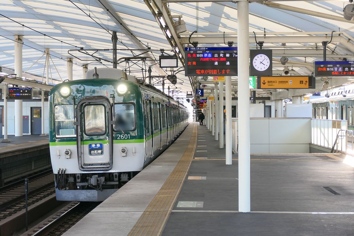 淀站高架月台,一列綠白色的京阪 2600 系電車進站,車頭方向牌是「準急 出町柳」;月台電子看板顯示先發 16:09 準急 出町柳、丹波橋で特急に連絡,次發「通過」,右側另一塊看板是 16:16 普通 出町柳;司機臉部已模糊