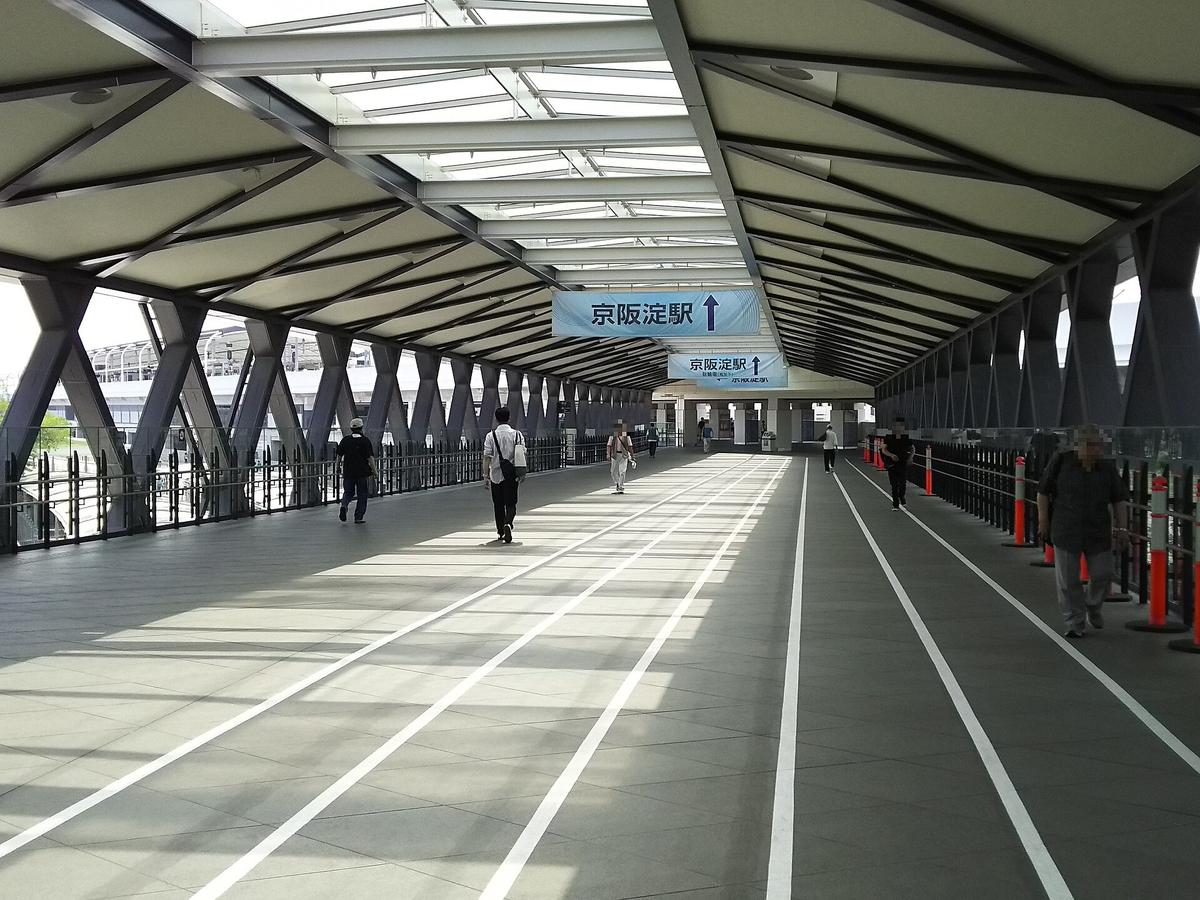 連接京都競馬場與淀站的寬闊有頂天橋,地面畫著白色分道線,吊牌寫「京阪淀駅 ↑」,只有零星幾位行人,部分行人臉部已由上傳者打上馬賽克