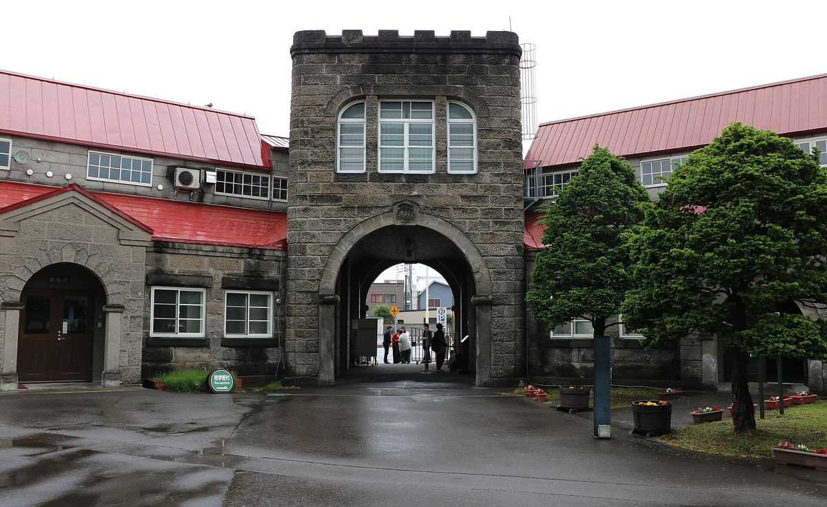 從余市蒸餾所廠區內側看正門:石砌塔樓下的拱門通往門外街道,兩側是紅色屋頂的石造建築,左下草叢邊有一塊綠色圓牌寫著「見学受付 Reception」與箭頭,雨後地面積水