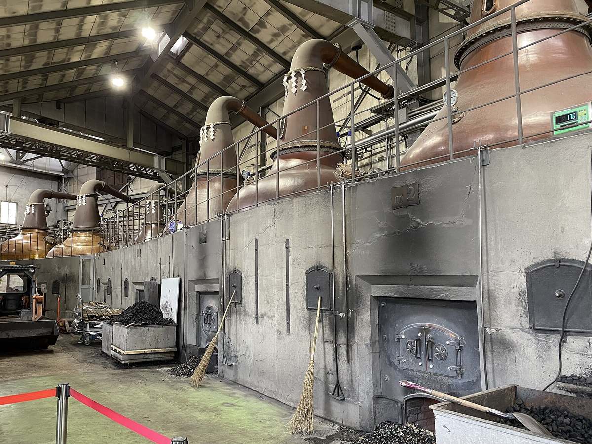余市蒸餾所蒸餾棟內部:水泥爐台上一排銅製蒸餾器,細長的頸部各綁著注連繩與白色紙垂,爐台正面是鑄鐵爐門,地上堆著煤炭、靠著兩支竹掃把,前景有紅色的參觀圍欄帶