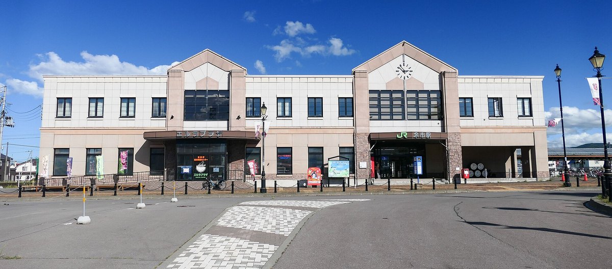 JR 余市站站舍正面:兩層樓的米白色建築,右側山牆上有時鐘、入口上方掛著「JR 余市駅」招牌,左側入口寫著「エルラプラザ」,站前是空曠的柏油廣場,藍天