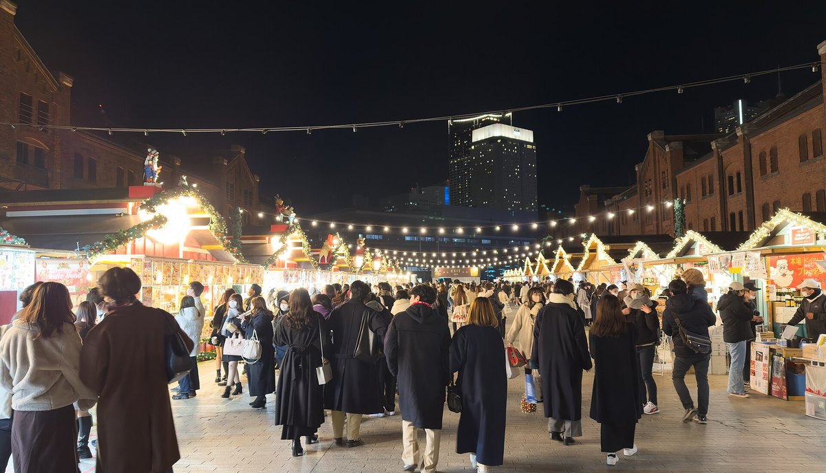 2022 年 12 月橫濱紅磚倉庫聖誕市集的攤位通道夜景：兩棟紅磚倉庫之間的石板廣場上，右側是一排屋簷纏著暖白燈串的三角屋頂木屋，左側是紅色屋頂、貼滿餐點照片的餐飲攤，頭頂橫拉著幾條燈泡串，中間是背對鏡頭往前走的人群