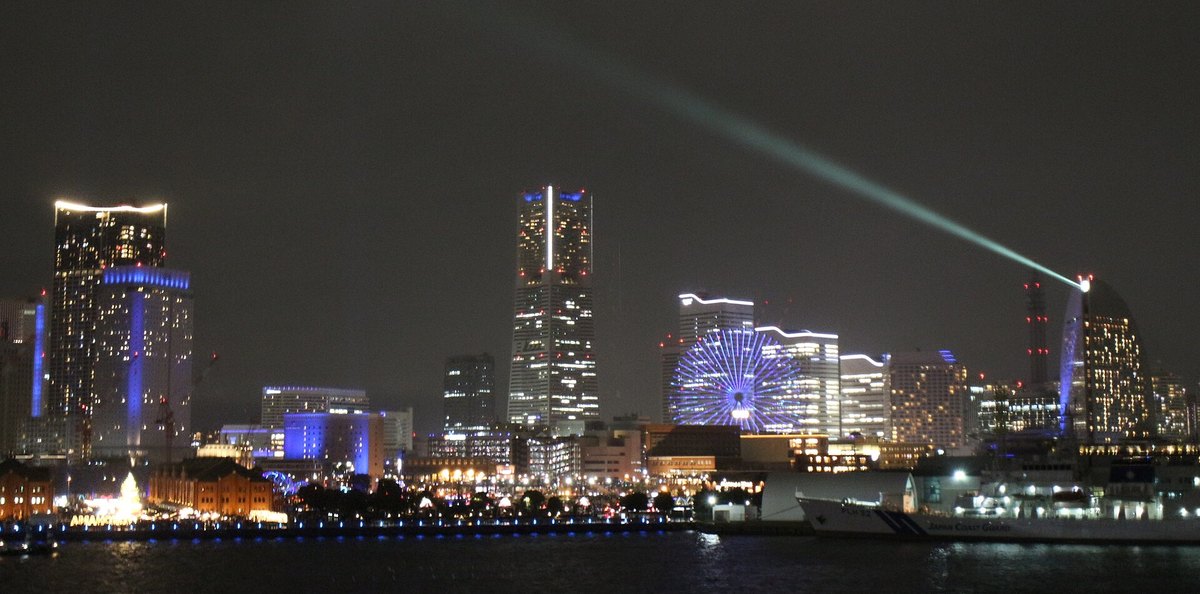 2021 年 12 月 25 日晚上從大棧橋看港未來：地標塔、摩天輪與海邊的大樓打著藍色燈光，右側帆船造型的飯店樓頂射出一道探照燈光束劃過夜空；左下角看得到兩棟紅磚倉庫與中間亮著的聖誕樹，右下角停著一艘白色的海上保安廳巡視船
