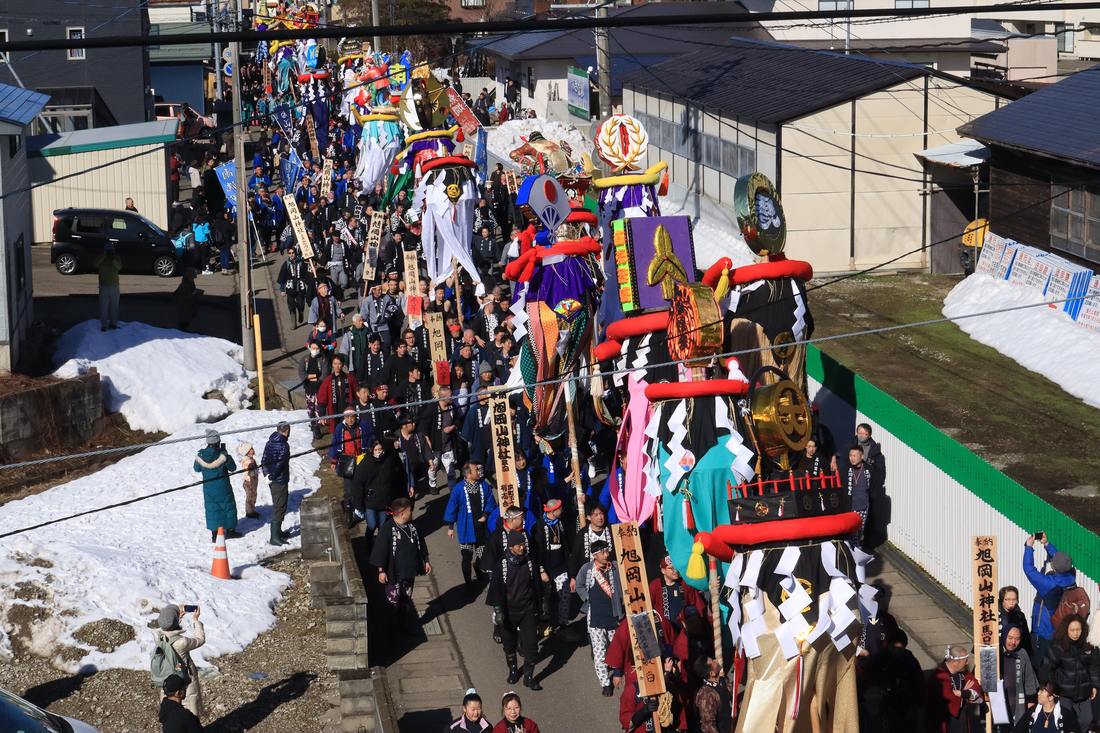 A Bonten column seen from a railway overbridge: a long line of poles topped with red-wrapped cylinders and black-and-white cloth skirts moving up a narrow street banked with leftover snow, wooden placards dedicating them to Asahiokayama Shrine held among the marchers