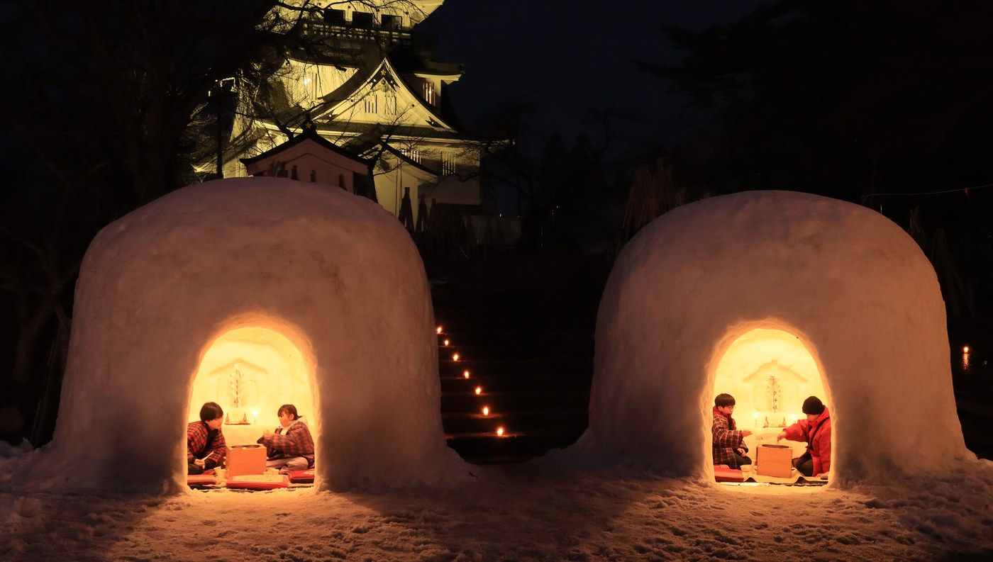 Warm light spilling from the arched openings of two snow domes, a small white altar with lit candles carved into the back wall of each, children seated on red cushions inside, a candle-lined snow staircase between them and the floodlit Yokote Castle behind