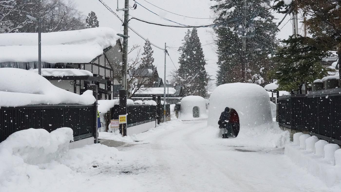 Heavy snow falling on the Haguro-cho samurai-house street in Yokote: a narrow cleared lane between black board fences and deeply snow-capped roofs, with two snow domes beside it and a visitor crouching at one entrance