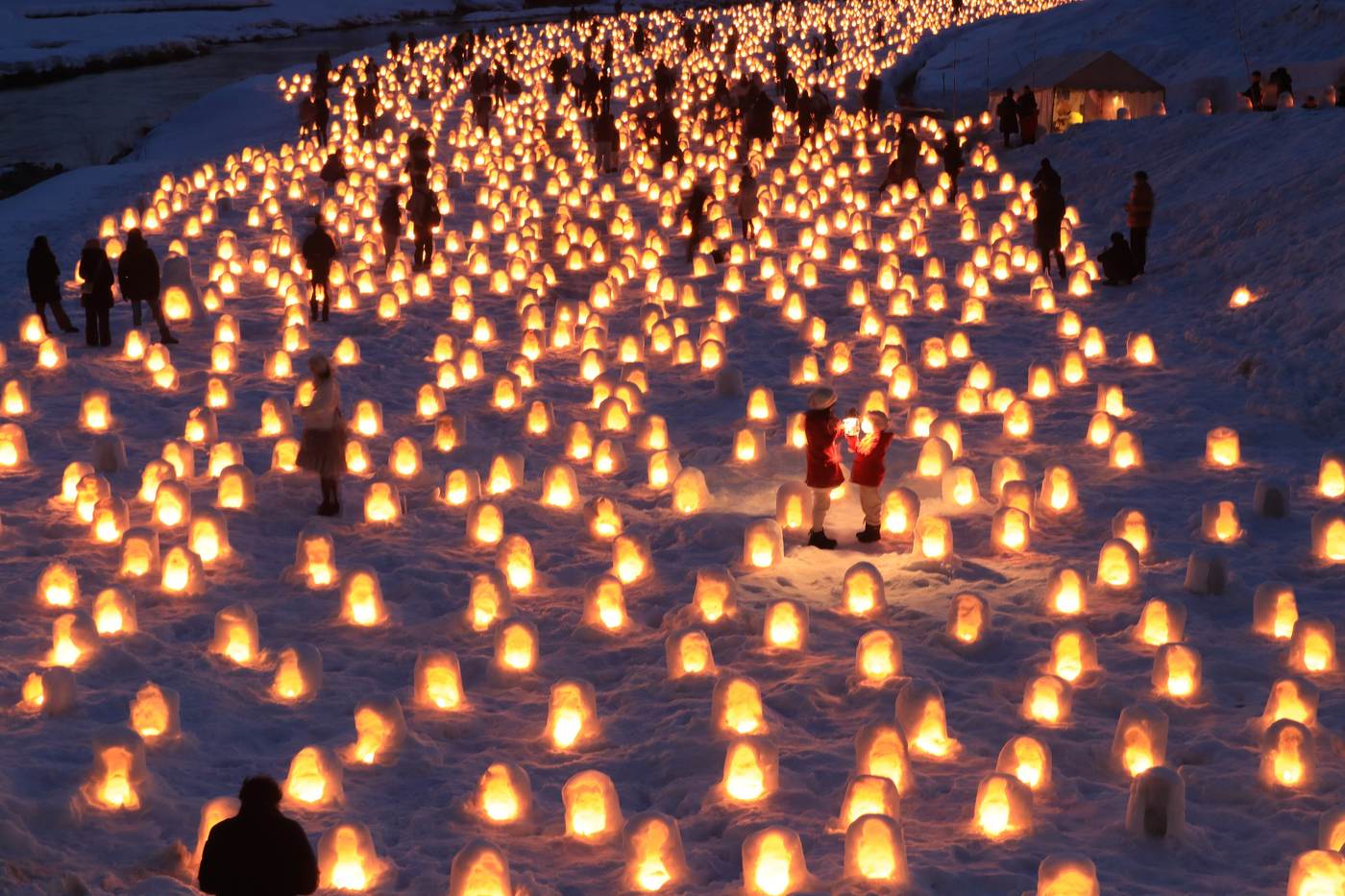 Janosaki Kawara seen from the bridge above: thousands of small candle-lit snow domes packed across the snowfield, the lights curving away along the riverbank with visitors scattered among them