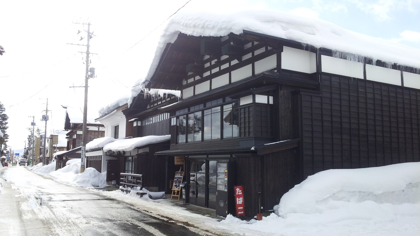 Winter on Nakananoka-machi street in Yokote: a black timber merchant house under a thick cap of roof snow with long icicles along the eaves, the roadway scraped to wet black asphalt and snow banked halfway up the shopfronts