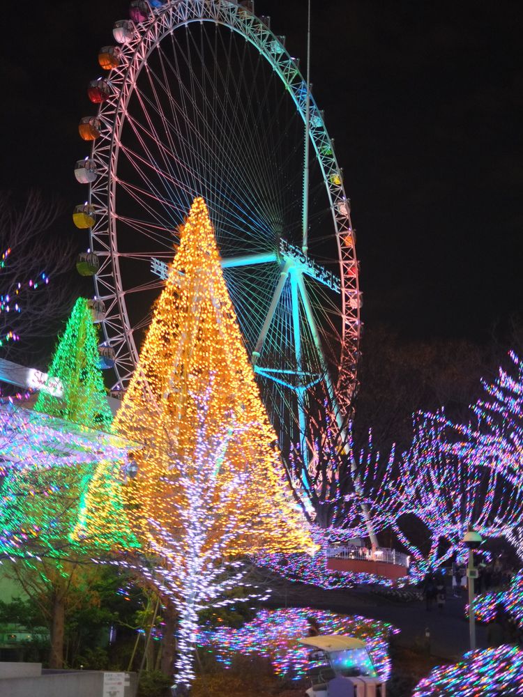 Yomiuriland at night: gold and green cone-shaped light trees in front of a large Ferris wheel with colored gondolas, foreground trees wrapped in pink, purple and white lights
