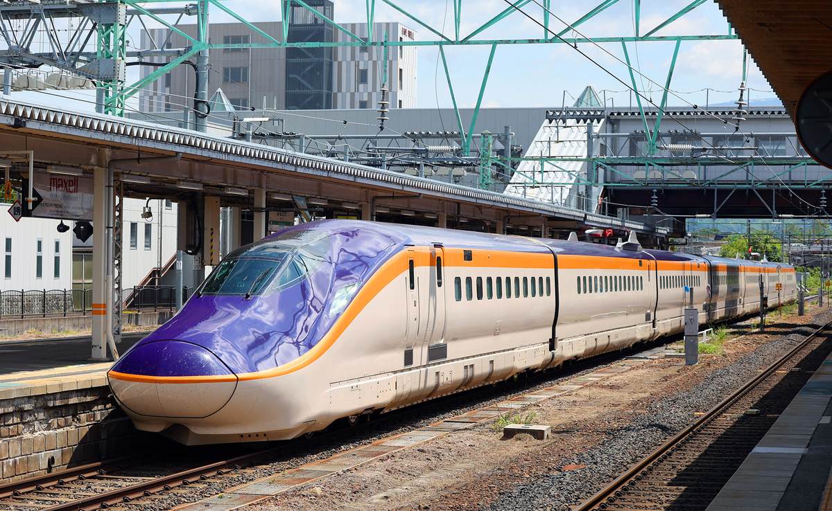 停在米澤站月台旁的山形新幹線 E8 系つばさ：車頭紫色、車身米白色配一條橘線，後方是綠色鋼架的跨線設施，晴天、地上沒有雪