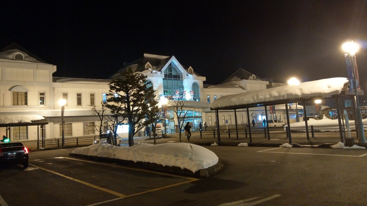 晚上的 JR 米澤站西口：白色站舍正面亮著「米沢駅」招牌，站前停車場邊堆著半人高的雪堆，右邊有頂棚積著厚雪的步道，幾個人影在站前走動
