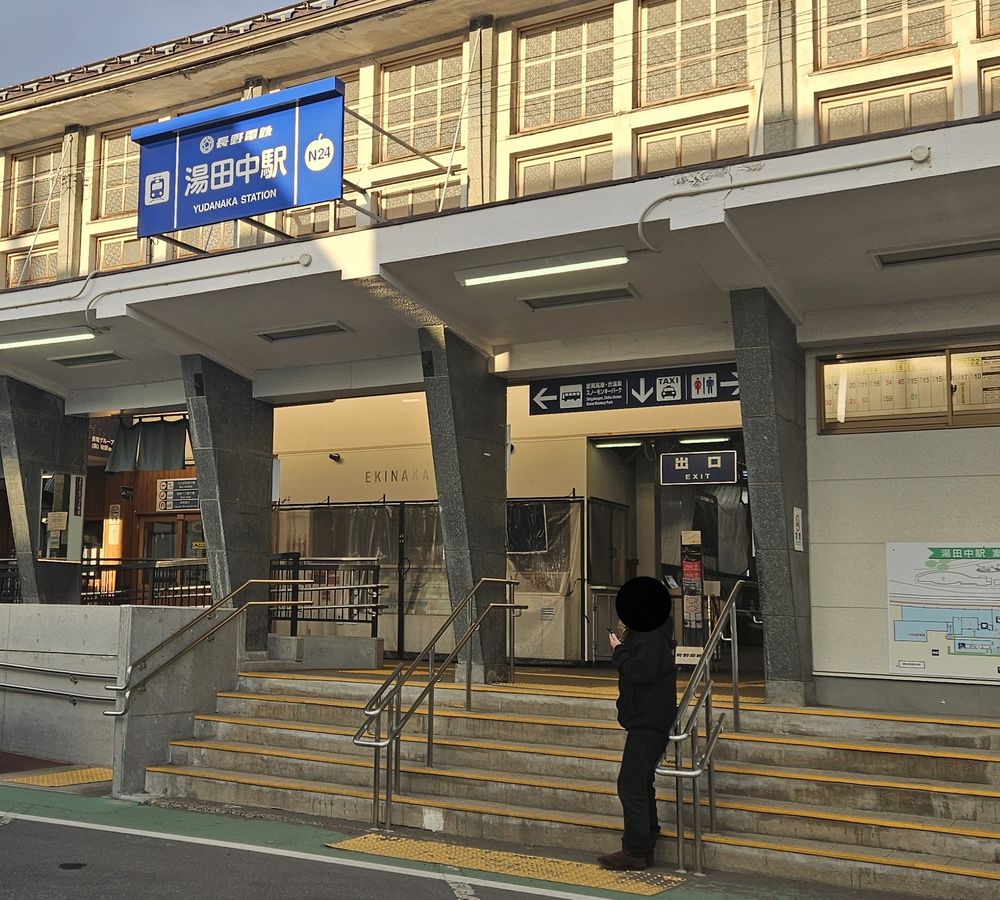 長野電鐵湯田中站正面，藍色站名牌「湯田中駅 N24」，入口上方的指標寫「志賀高原・渋温泉 スノーモンキーパーク」並有巴士圖示與往左的箭頭