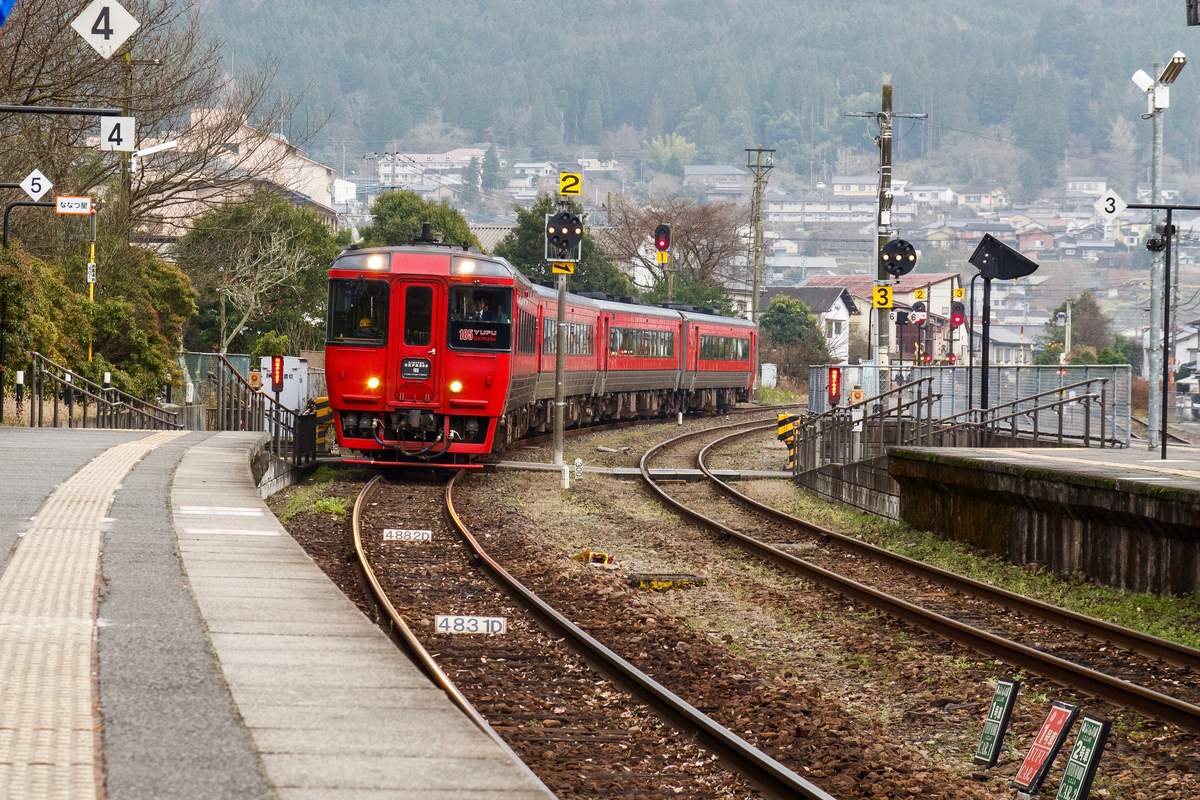 紅色的特急由布號キハ185 系柴油車駛進由布院站,車頭標示 185 YUFU,背景是冬天的由布院市街與山坡