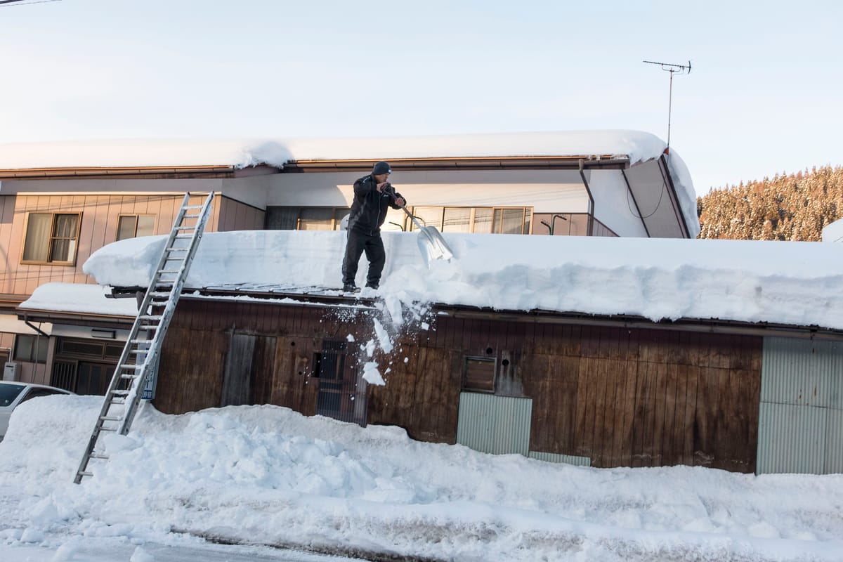 Yukioroshi in a snow-country residential street: a resident on a ladder shovels a metre-thick layer of snow off a roof into the road below