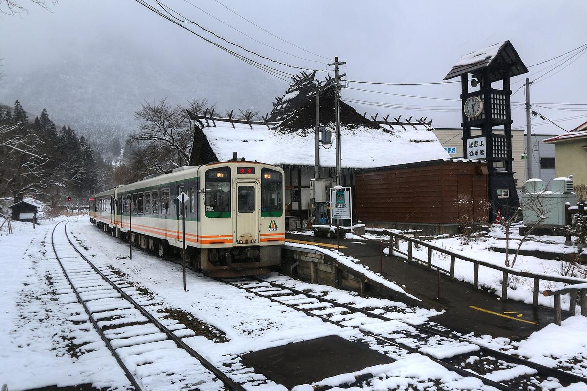 下雪的湯野上溫泉站：一列米白色車身、橘綠腰線的會津鐵道列車停在月台邊，車頭顯示「快速」，後方是積雪的茅葺屋頂站舍與寫著站名的木造鐘塔