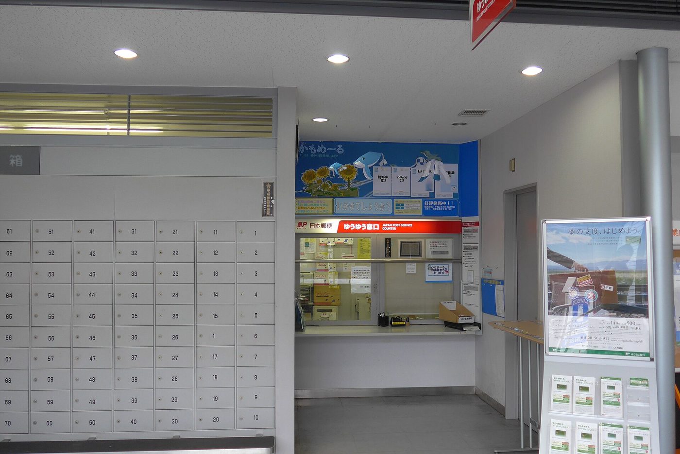 A small service window inside a Japanese post office with a red sign reading Yu-yu Madoguchi, Japan Post Service Counter, alongside the Japan Post logo; brown cartons and white parcels are stacked on the counter shelf inside, notices are taped to the wall, a full wall of numbered post office boxes from 1 to 70 fills the left half of the frame, and a Japan Post Bank standing display board is on the right
