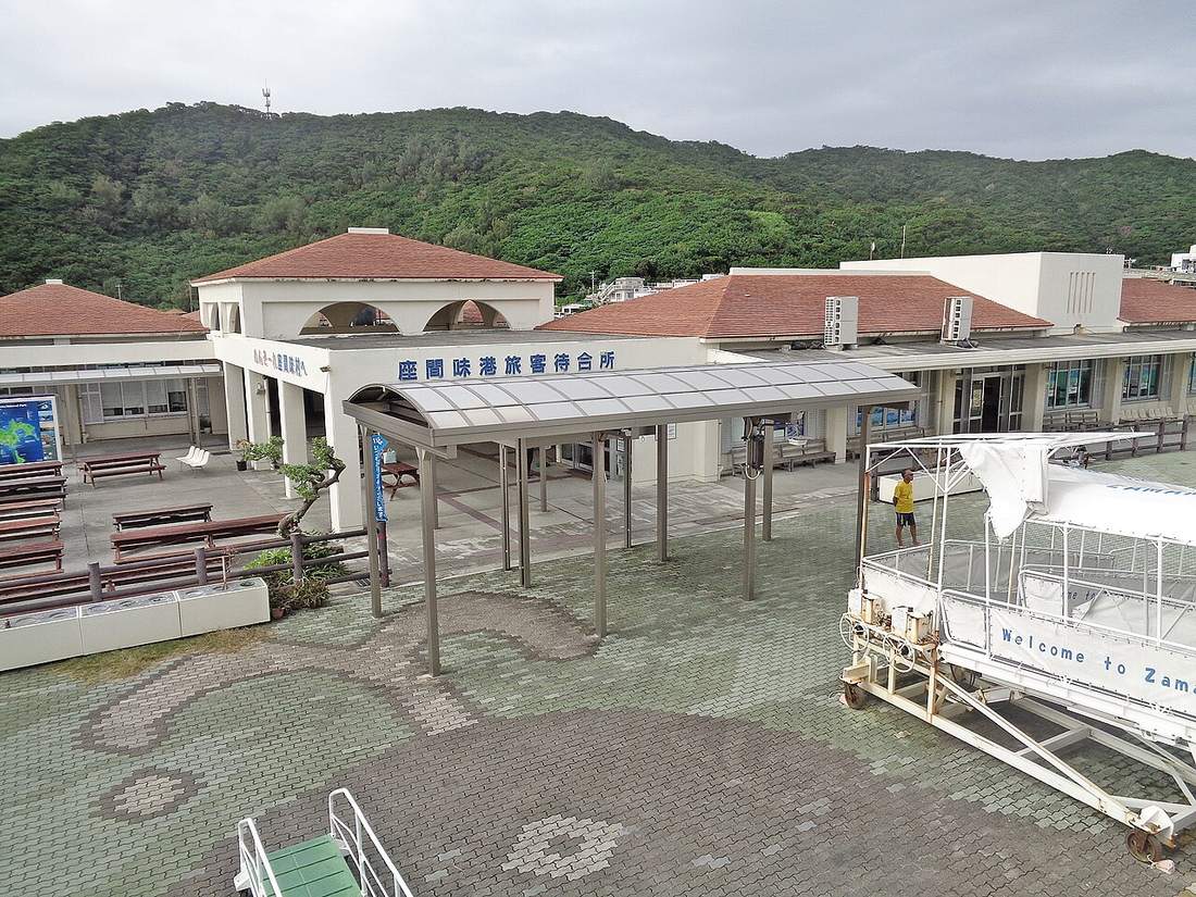 The exterior of Zamami Port's passenger waiting hall, a low red-tiled building with its Japanese name in blue lettering across the front, empty benches and an arched canopy in front, a wheeled boarding gangway marked Welcome to Zamami at right, green hills and overcast sky behind