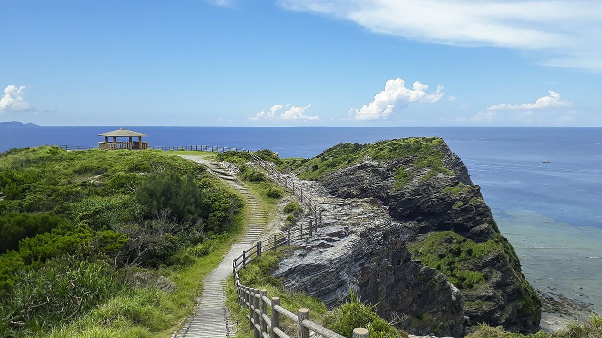 The Unajinosachi observatory on Zamami, a wooden hexagonal shelter on a grassy headland with a railed path climbing towards it along a rocky cape, open deep blue sea to the right and island silhouettes on the horizon