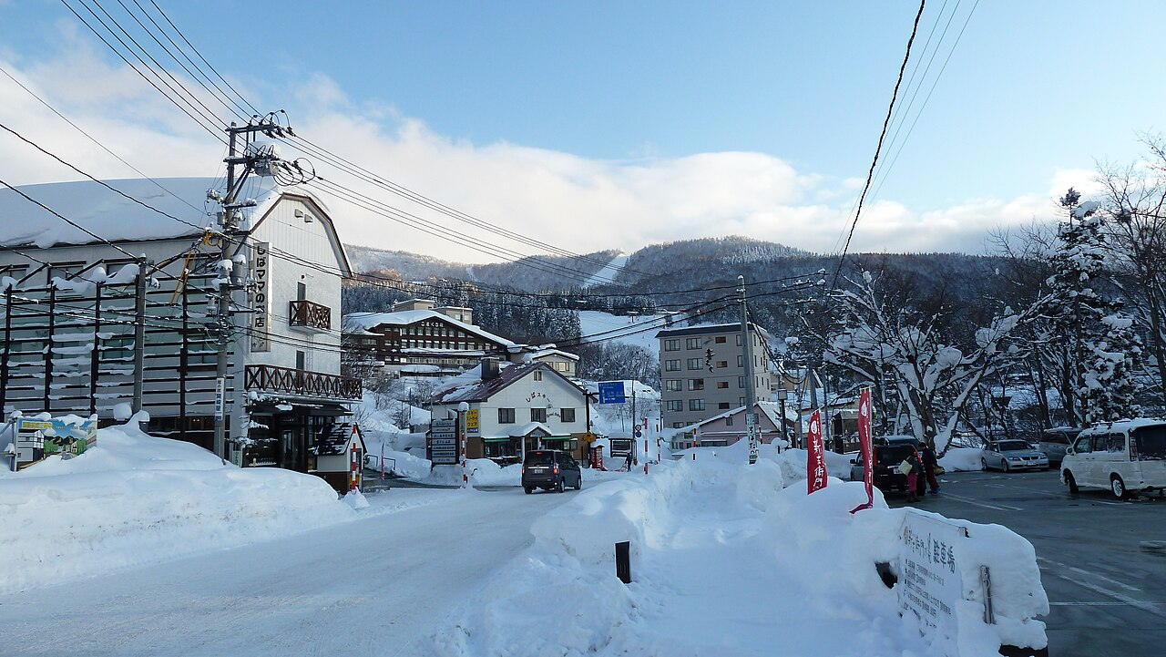 Winter street in Zao Onsen: packed snow road walled in by waist-high snowbanks, timber inns and lodges lining both sides, ropeway cables crossing overhead, snow-covered slopes behind