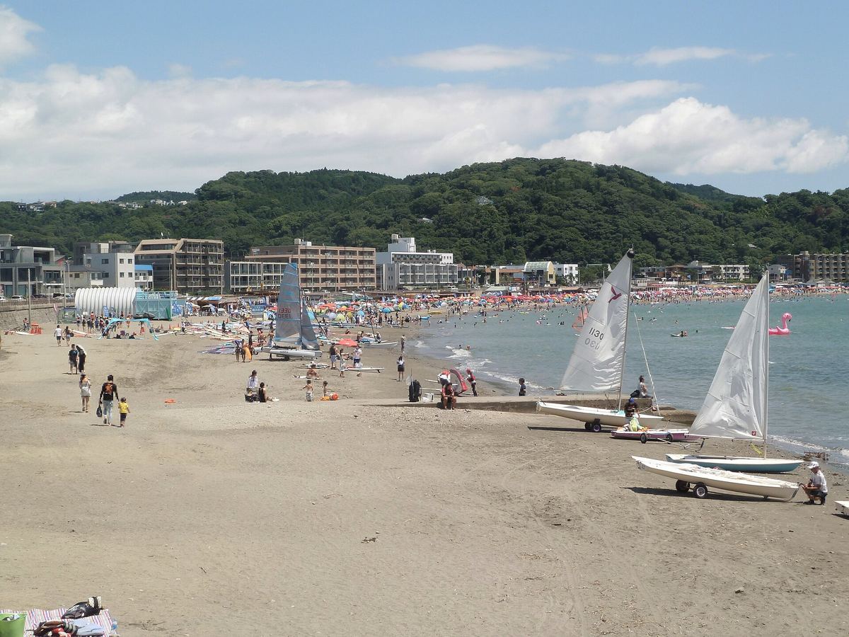Zushi Beach in summer: an empty stretch of brown sand in the foreground, a long line of beach-house tents and coloured parasols running along the shore behind it with a scattering of distant bathers, three sailing dinghies parked on the sand to the right, and low green hills with white apartment blocks around the bay